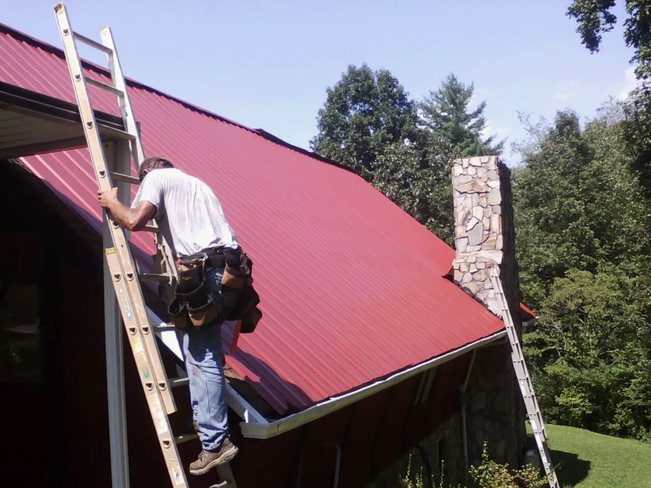 Roofer on a ladder beside a red metal roof and stone chimney — Soffit & Fascia Repair by Roofing Brothers in South Hadley